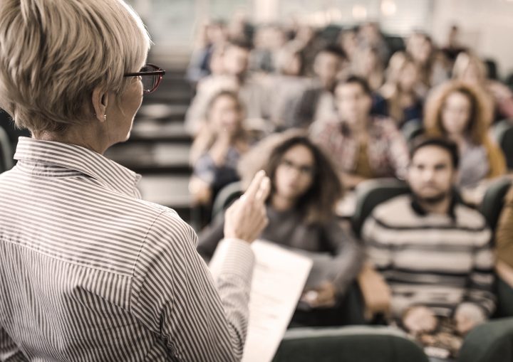 Back view of a senior professor talking on a class to large group of students.