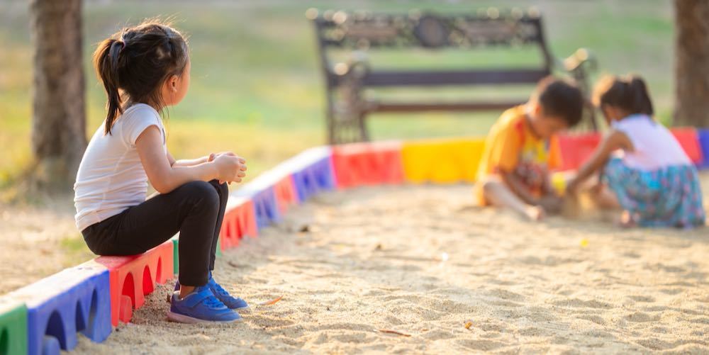 Little girl sitting lonely watching friends play at the playground.The feeling was overlooked by other people.Concept child shy.