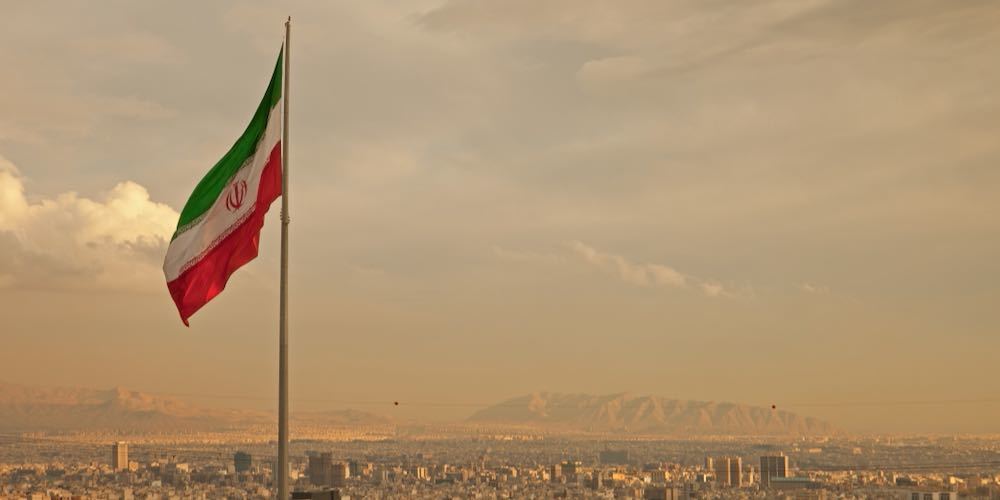 Iran flag waving  in the wind above skyline of Tehran lit by orange glow of sunset.