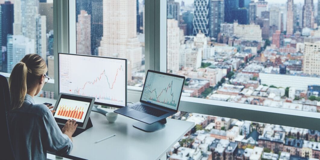 Back view of business woman sitting at panoramic skyscraper office desktop front PC computer with financial graphs and statistics on monitor.Analysis of digital market and investment in block chain