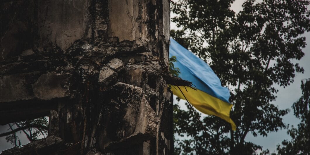 A Ukrainian flag waves in the background of war torn ruins, captured with an artistic blur effect. The image symbolizes resilience amidst destruction during the war in Ukraine