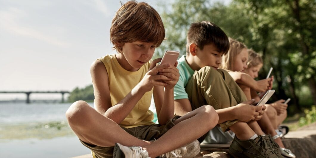 Row of multiethnic children using and browsing mobile phones on log on river coast outdoors. Boys and girl of generation alpha. Gadget addiction. Childhood lifestyle. Sunny summer day